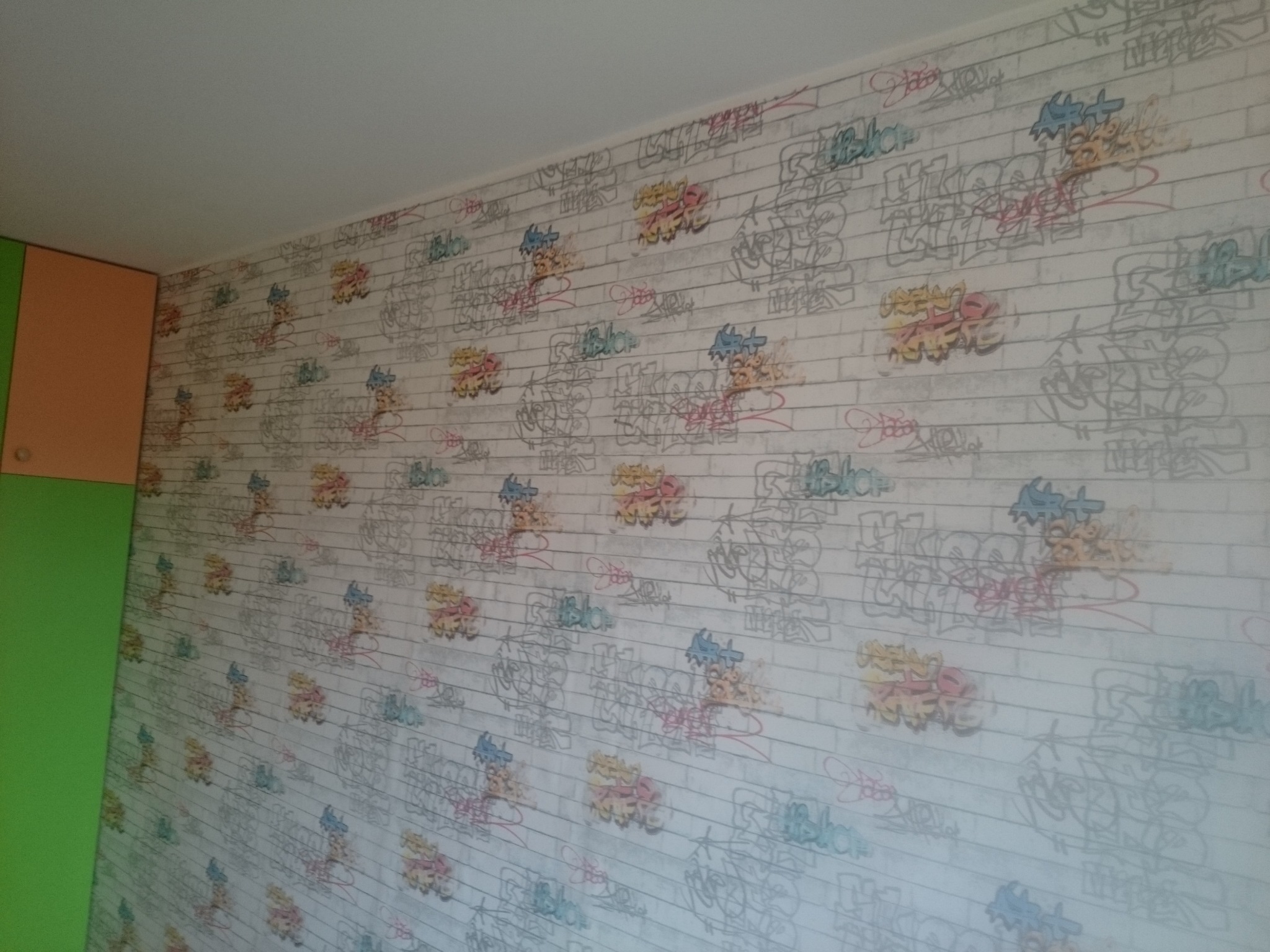 Ściana pokryta tapetą w stylu graffiti, z motywem cegieł i kolorowymi napisami, przylegająca do zielonej i pomarańczowej szafki.