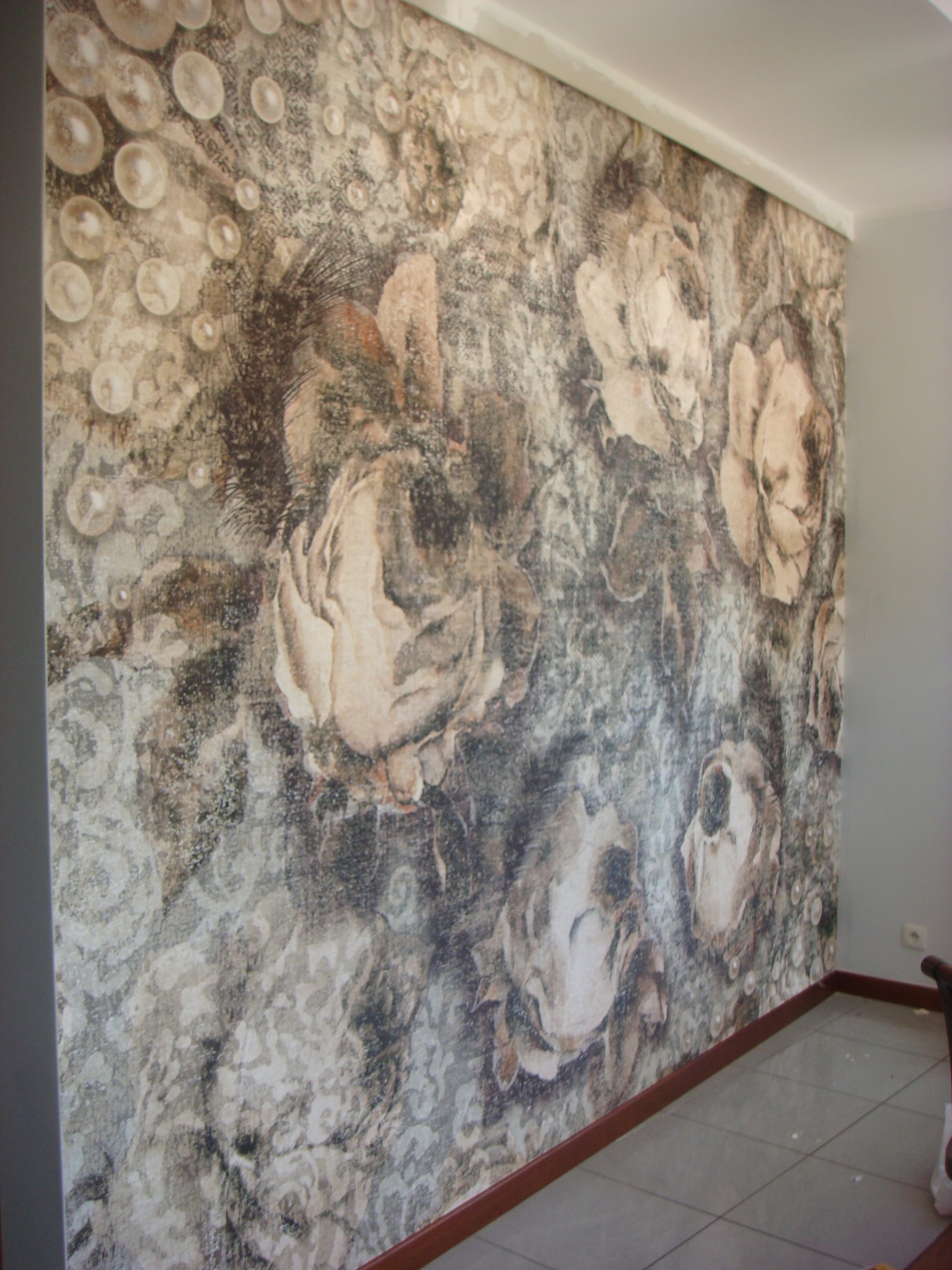 Ściana po tapetowaniu z wzorem kwiatów i bąbelków w odcieniach szarości i beżu, widoczny fragment podłogi z płytek.