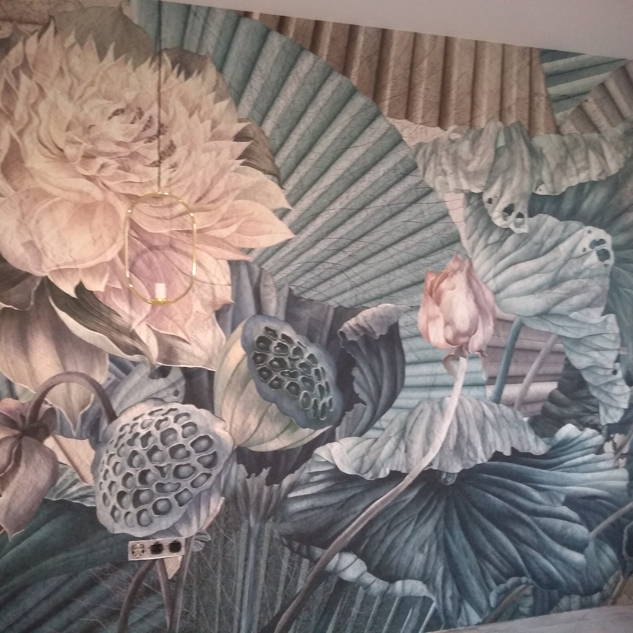 Ściana pokryta tapetą z motywem kwiatów lotosu i liści w odcieniach beżu, szarości i zieleni, z widoczną lampką nocną i gniazdkiem elektrycznym.