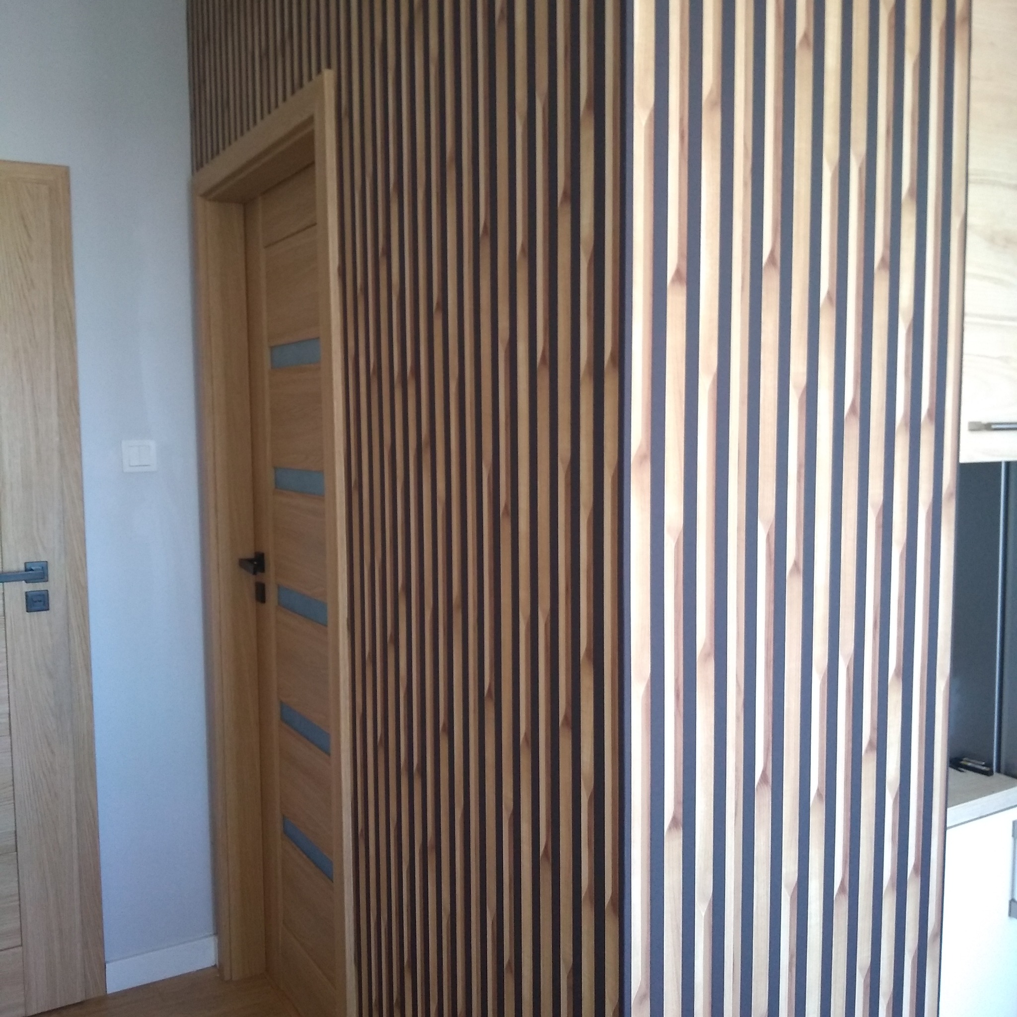 Drewniane lamele ścienne w odcieniach beżu i grafitu, tworzące nowoczesny wzór w pionowe pasy, widoczne obok drewnianych drzwi z przeszkleniami.