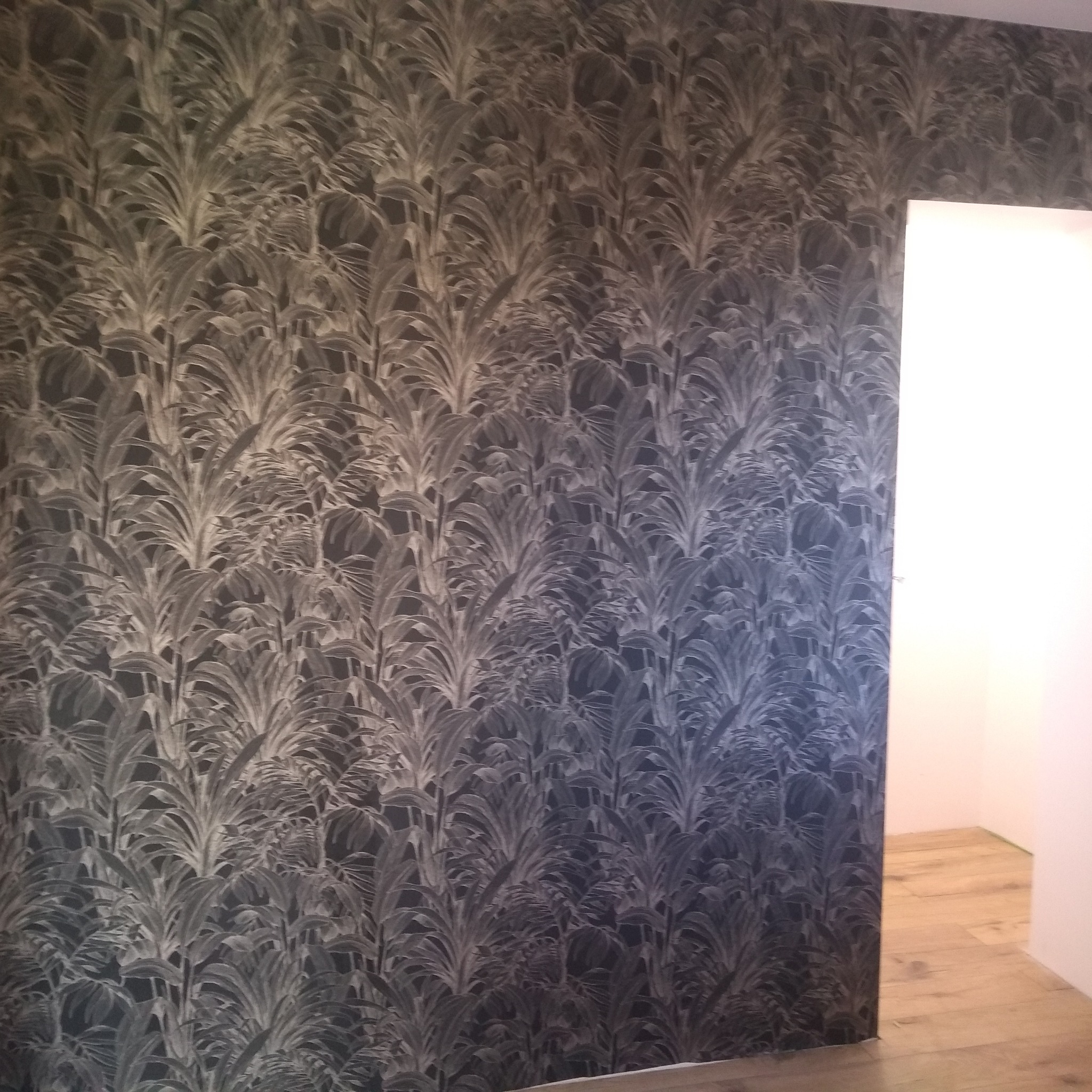 Ściana pokryta tapetą w ciemnych odcieniach z motywem roślinnym, widok na fragment podłogi i przejście do innego pomieszczenia.