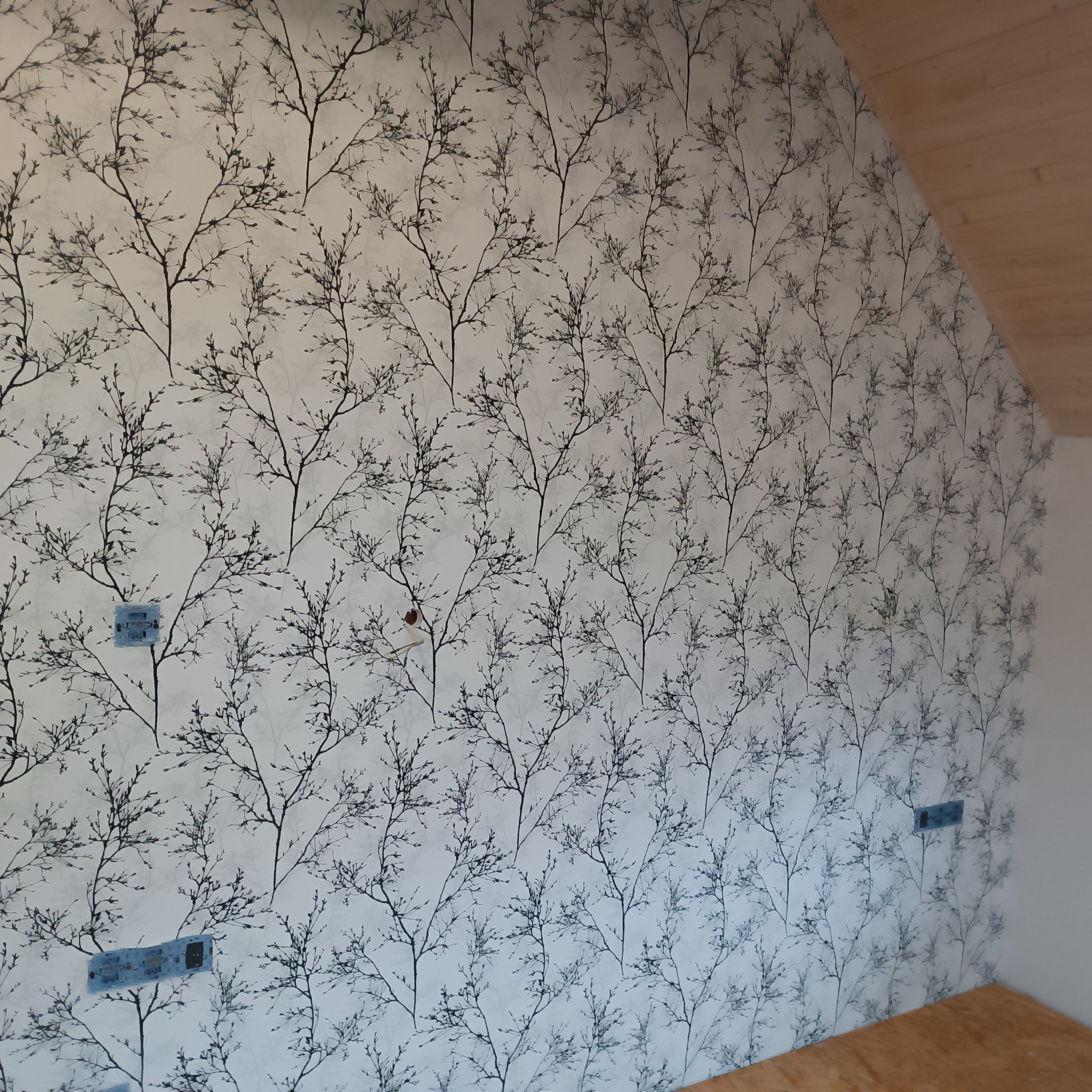 Ściana w pokoju na poddaszu pokryta tapetą z motywem gałęzi drzew na jasnym tle, widoczne gniazdka elektryczne.