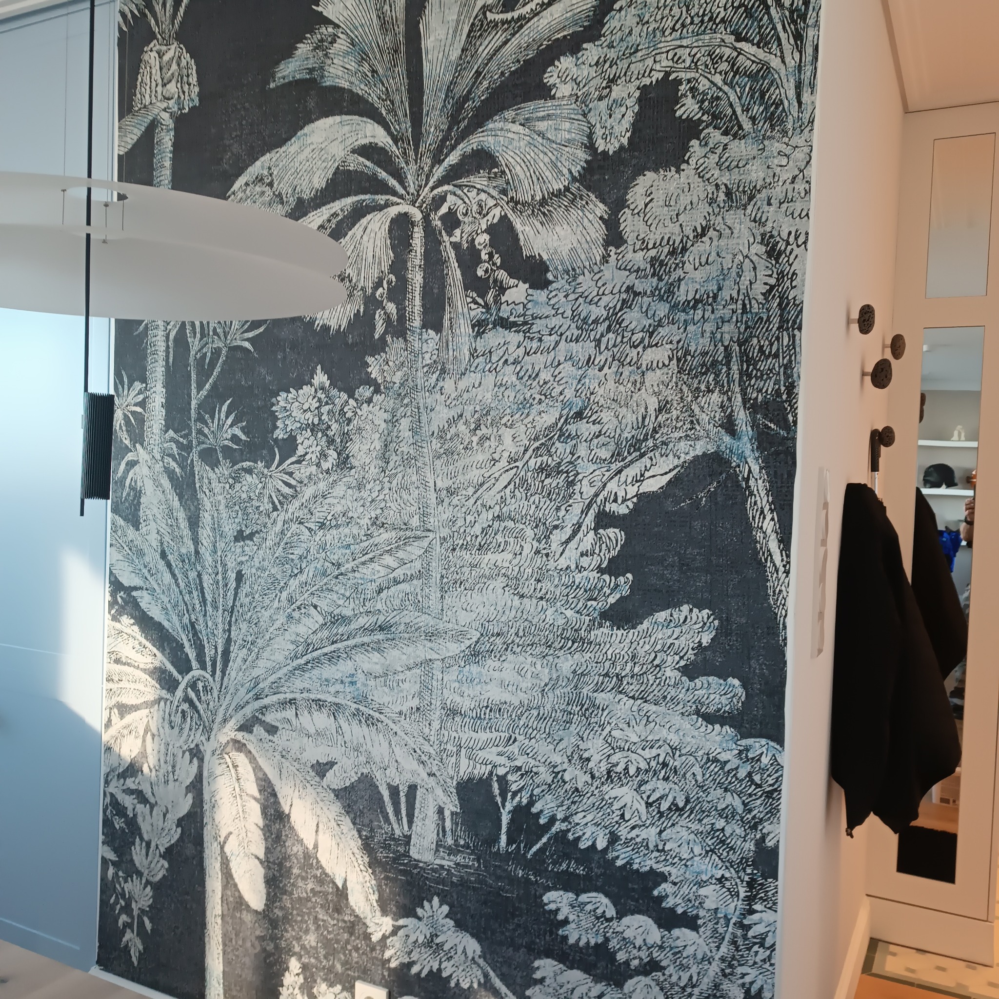 Fragment ściany z tapetą w stylu retro przedstawiającą tropikalny las w odcieniach bieli i granatu, z widoczną lampą wiszącą i wieszakiem na ubrania.