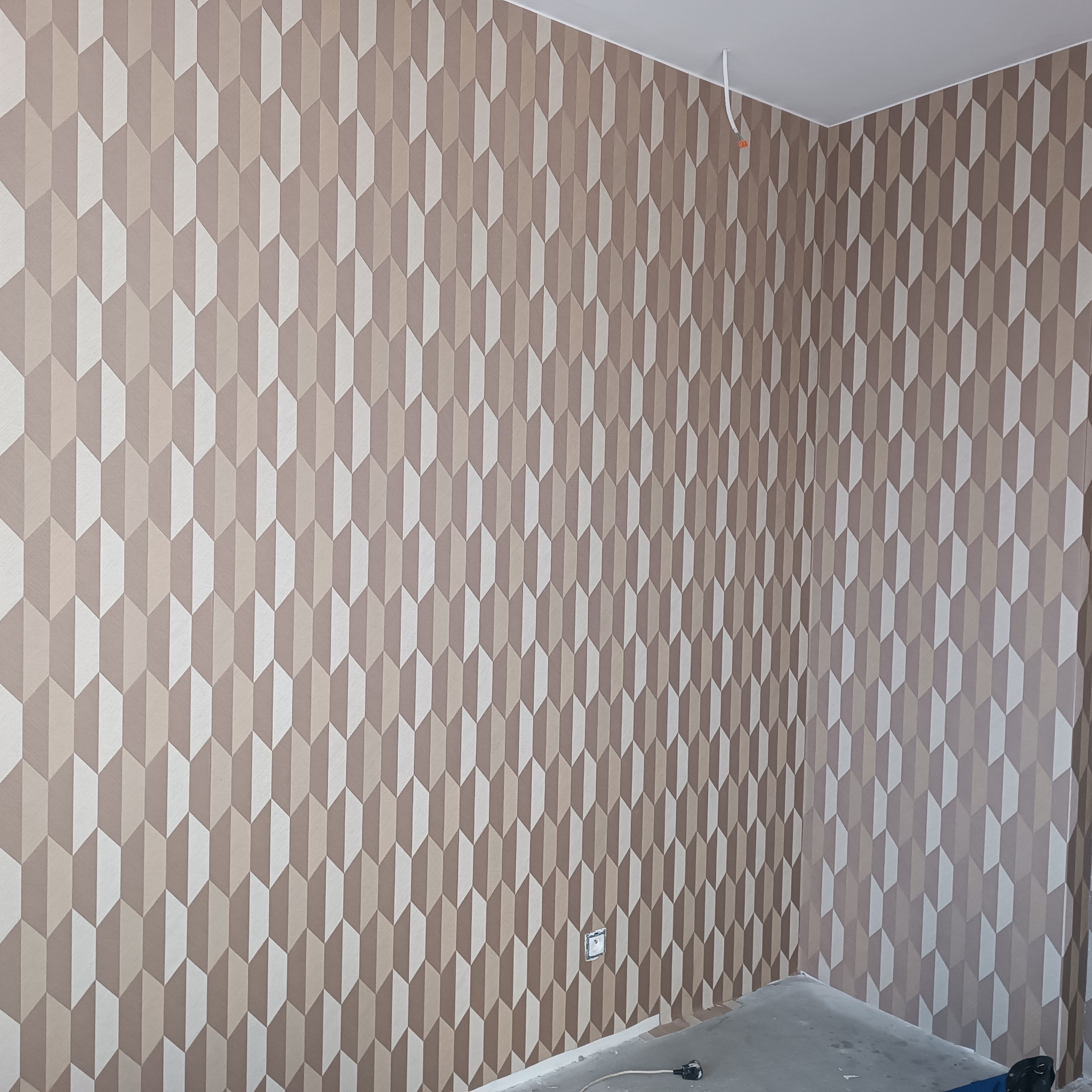 Tapeta w odcieniach beżu i bieli z geometrycznym wzorem strzałek, świeżo położona na ścianie w rogu pokoju, widoczny fragment podłogi i gniazdko elektryczne
