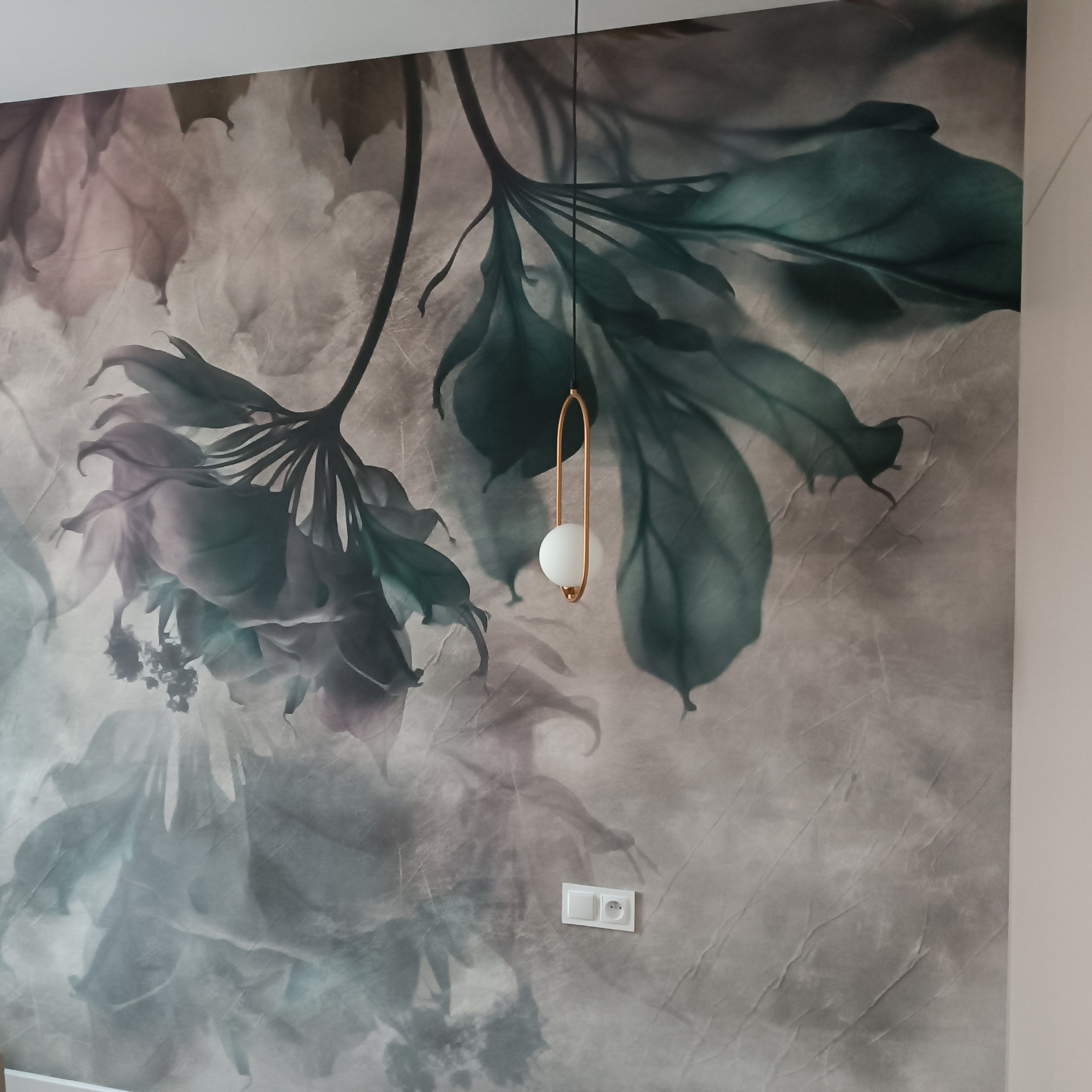 Ściana pokryta tapetą z motywem kwiatowym w odcieniach szarości, zieleni i różu, oświetlona wiszącą lampą z okrągłym kloszem i złotym stelażem, widoczny włącznik światła.