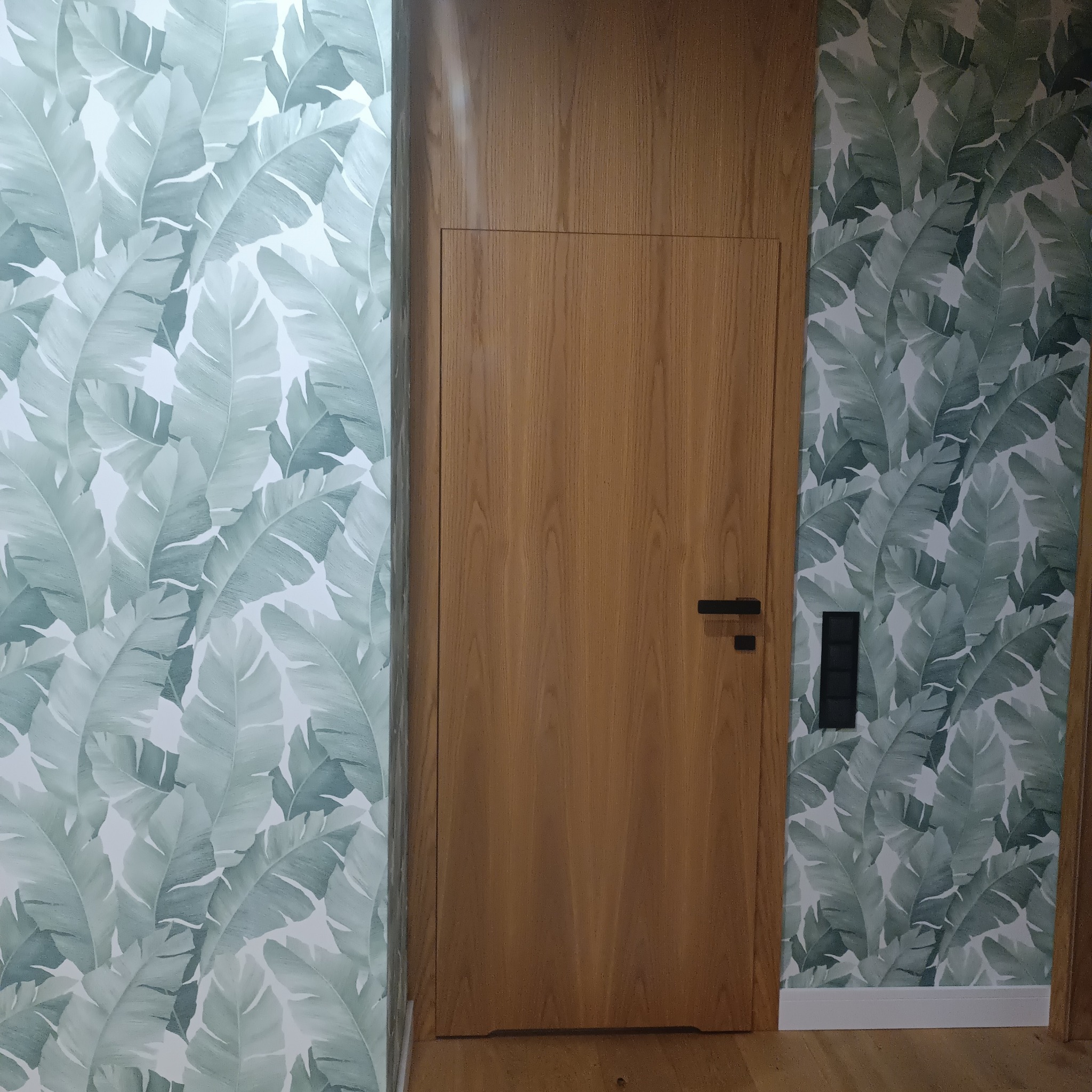 Zamknięte drewniane drzwi z czarną klamką, przylegające do ściany pokrytej tapetą w duże, zielono-białe liście.