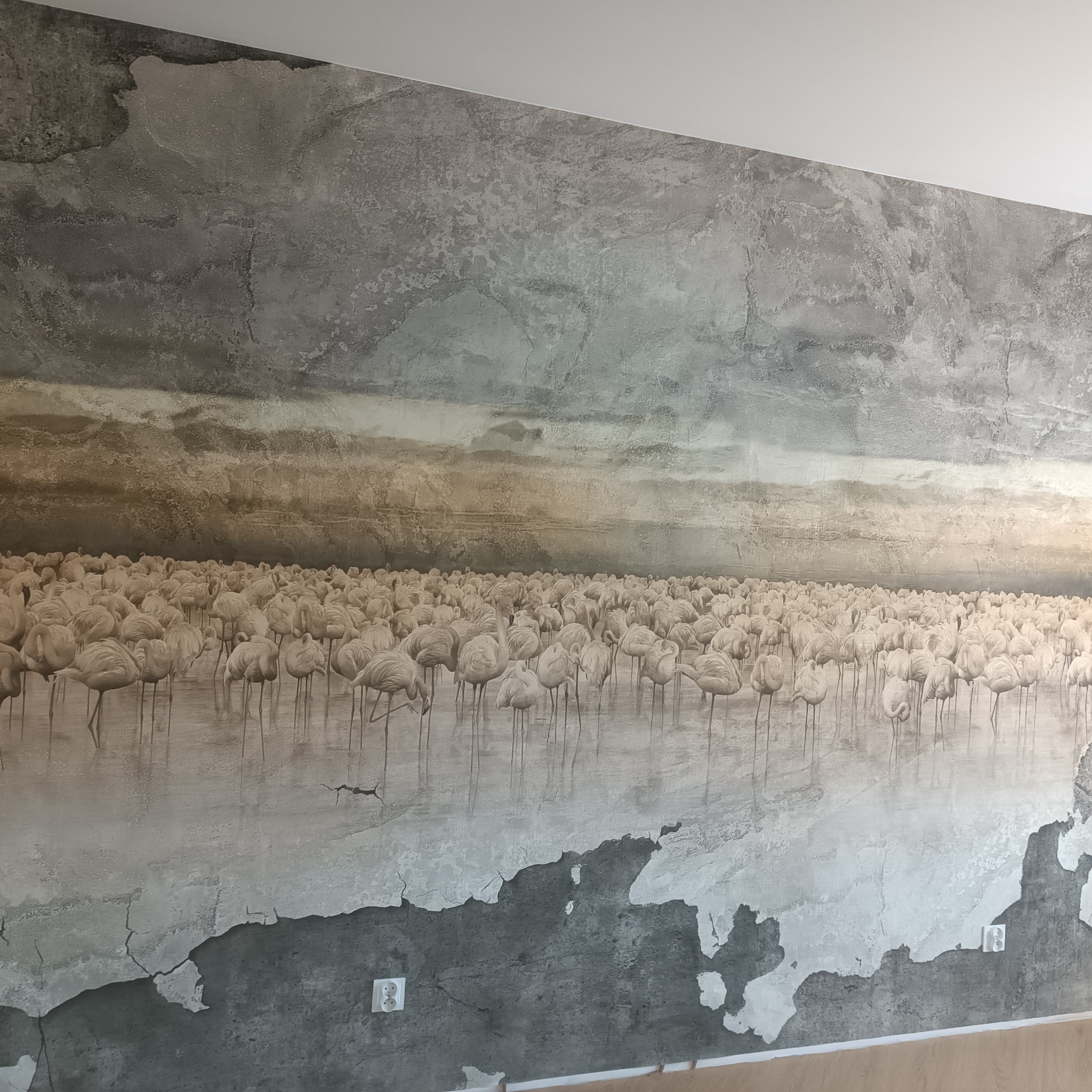 Ściana w Gdańsku z fototapetą przedstawiającą stado flamingów na tle pochmurnego nieba, imitująca efekt starego, popękanego tynku.