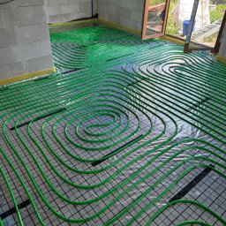 Izoinstal - Zielone rurki ogrzewania podłogowego ułożone spiralnie na czarnej siatce, widok z góry w surowym wnętrzu z oknem na ogród.