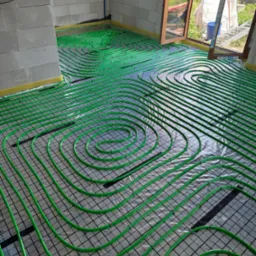 Zielone rurki ogrzewania podłogowego ułożone spiralnie na czarnej siatce, widok z g&oacute;ry w surowym wnętrzu z oknem na ogr&oacute;d.