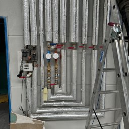 Izoinstal - Izolowane rury instalacji grzewczej z zaworami i licznikiem ciepła, drabina Krause oparta o ścianę, fragment płyty karton-gips w dolnym rogu.