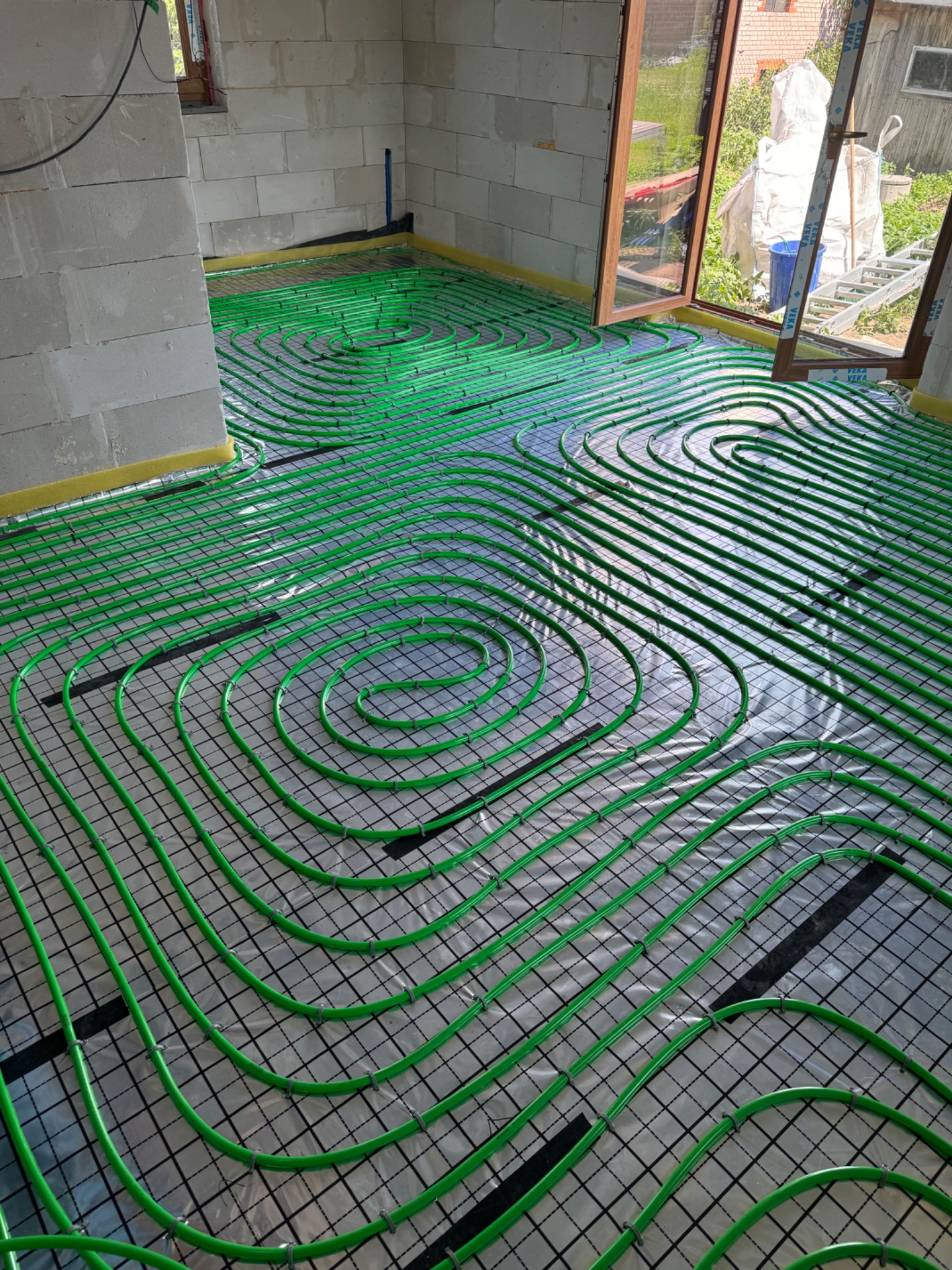 Zielone rurki ogrzewania podłogowego ułożone spiralnie na czarnej siatce, widok z góry w surowym wnętrzu z oknem na ogród.