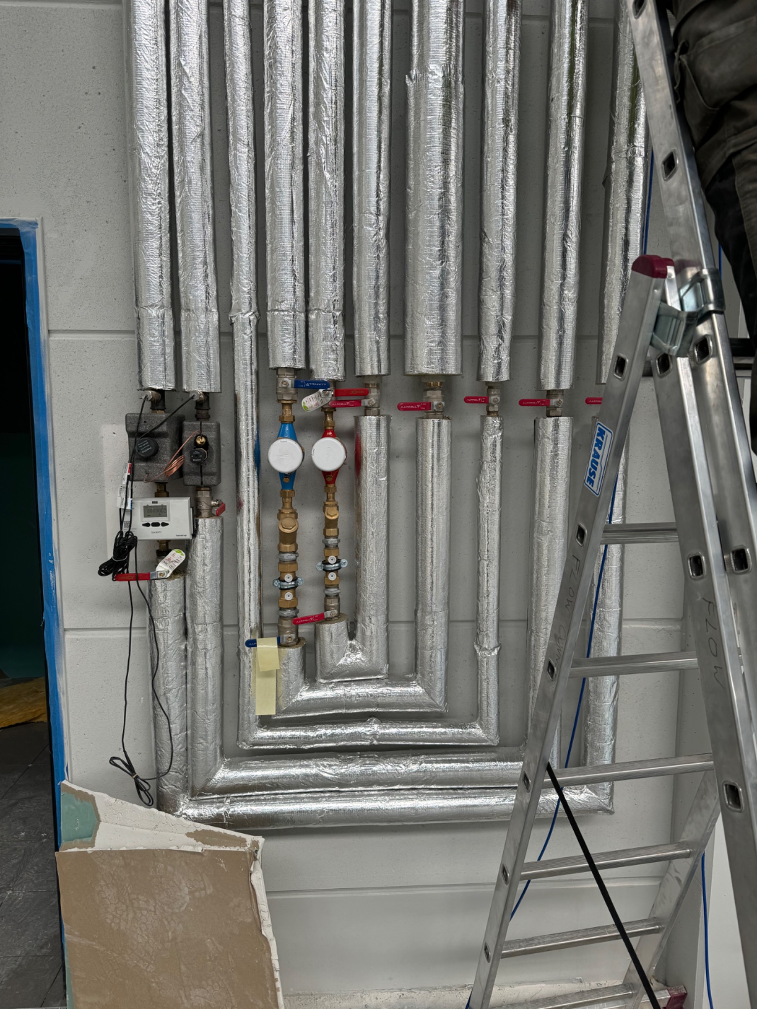 Izolowane rury instalacji grzewczej z zaworami i licznikiem ciepła, drabina Krause oparta o ścianę, fragment płyty karton-gips w dolnym rogu.