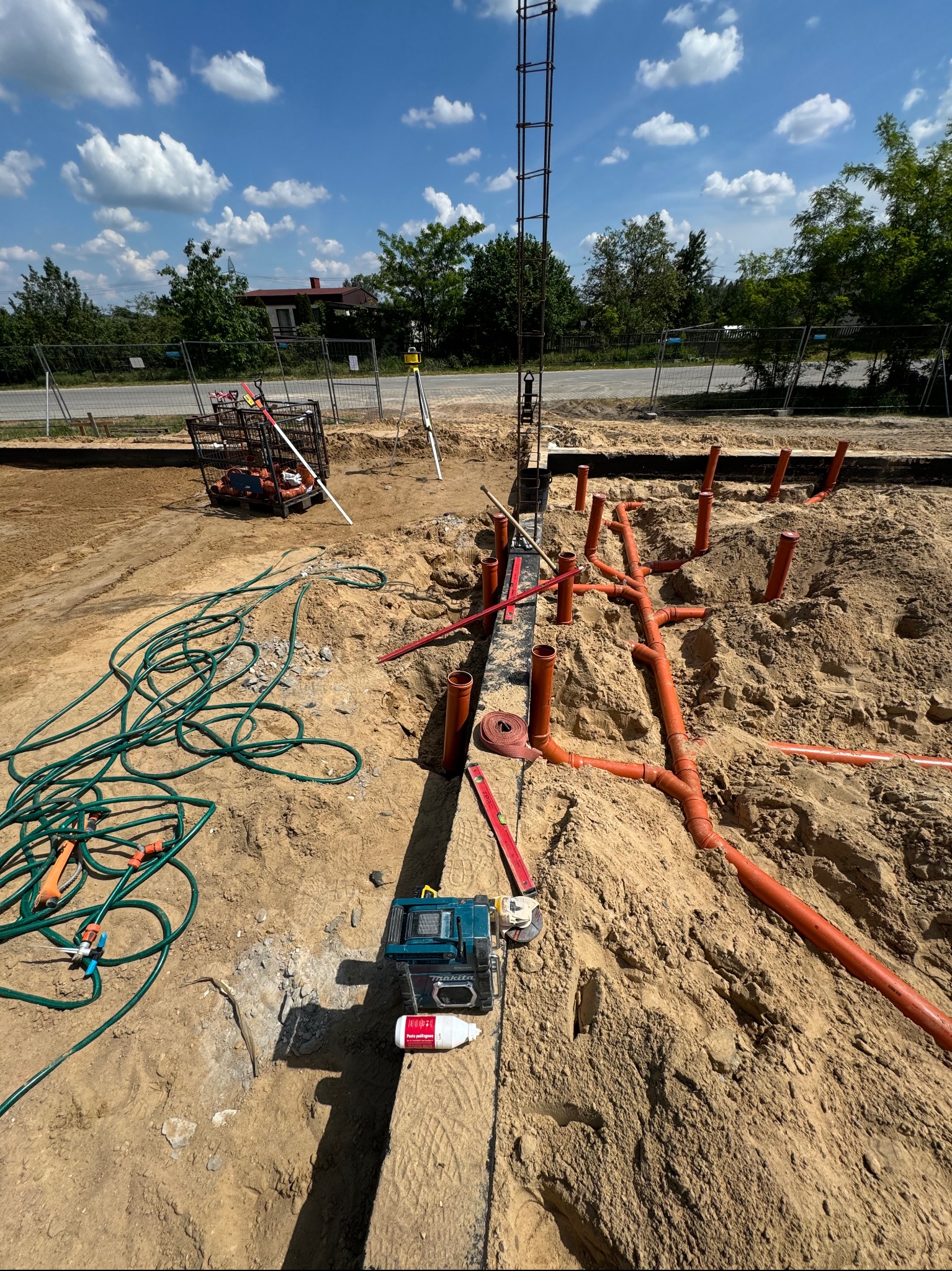 Rozłożone pomarańczowe rury kanalizacyjne w piasku na placu budowy, widoczny laserowy niwelator, szpula taśmy mierniczej i narzędzia.