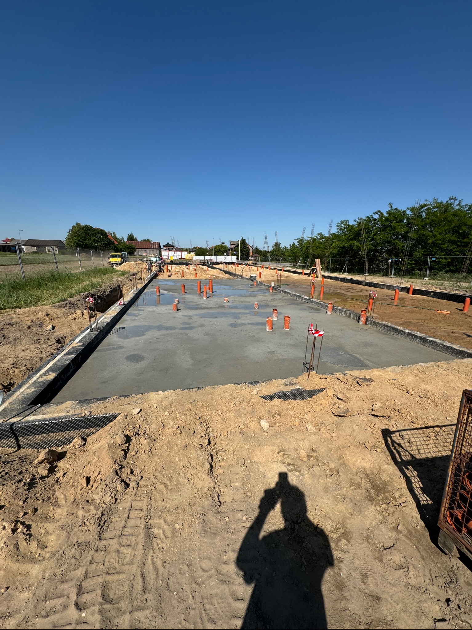 Wylany fundament z wystającymi rurami kanalizacyjnymi i elementami zbrojenia, widok z góry w słoneczny dzień z cieniem fotografa.