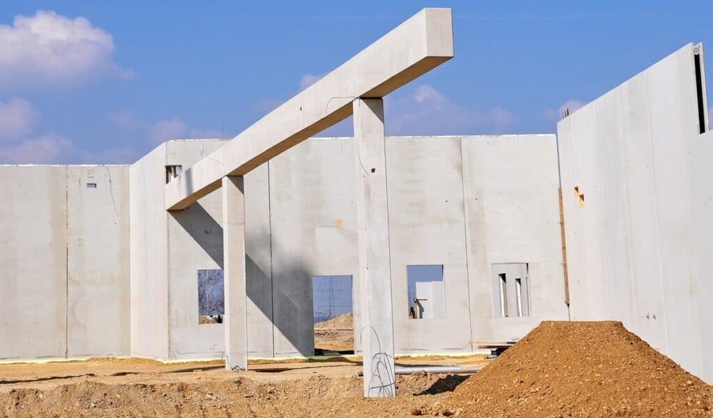 Surowy stan budowy: szkielet żelbetowy z kolumnami i belkami, widoczne otwory okienne, nasyp ziemi w tle, błękitne niebo.