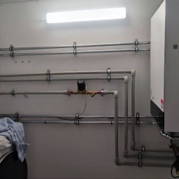 Duo Termika Paweł Tarkowski - Kompaktowa kotłownia gazowa z białym kotłem wiszącym Viessmann, niebieskim zbiornikiem wyrównawczym i licznymi rurami w małym, oświetlonym pomieszczeniu. Na pralce leżą narzędzia i ubrania.