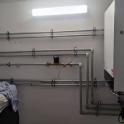 Kompaktowa kotłownia gazowa z białym kotłem wiszącym Viessmann, niebieskim zbiornikiem wyrównawczym i licznymi rurami w małym, oświetlonym pomieszczeniu. Na pralce leżą narzędzia i ubrania.