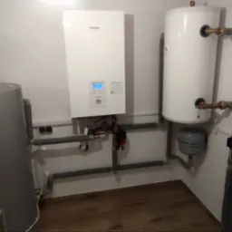 Instalacja grzewcza w kotłowni: pompa ciepła Panasonic, zasobnik Ferolli z niebieskim naczyniem wzbiorczym, biały zbiornik buforowy i szare rury instalacyjne.