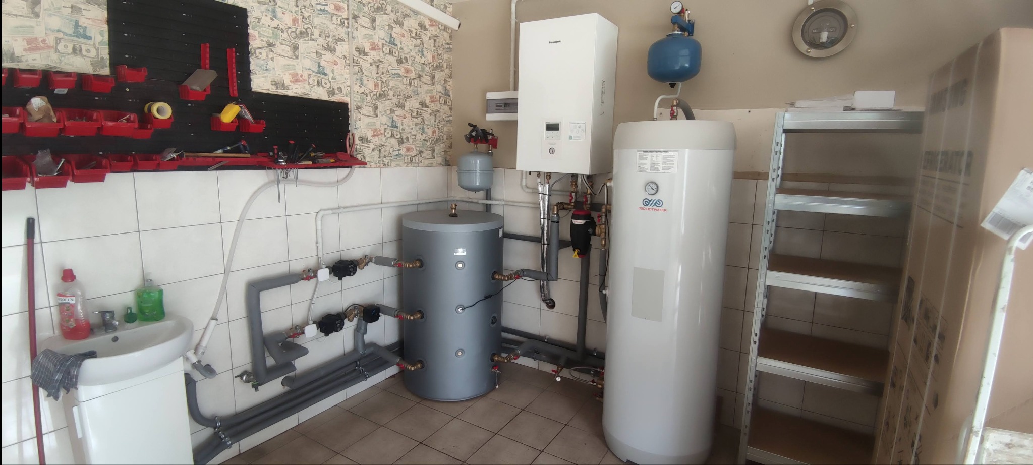 Zainstalowana pompa ciepła powietrze-woda Panasonic z szarym zasobnikiem, widoczna instalacja hydrauliczna z rurami w izolacji i elementami sterującymi, na ścianie tablica narzędziowa z czerwonymi...