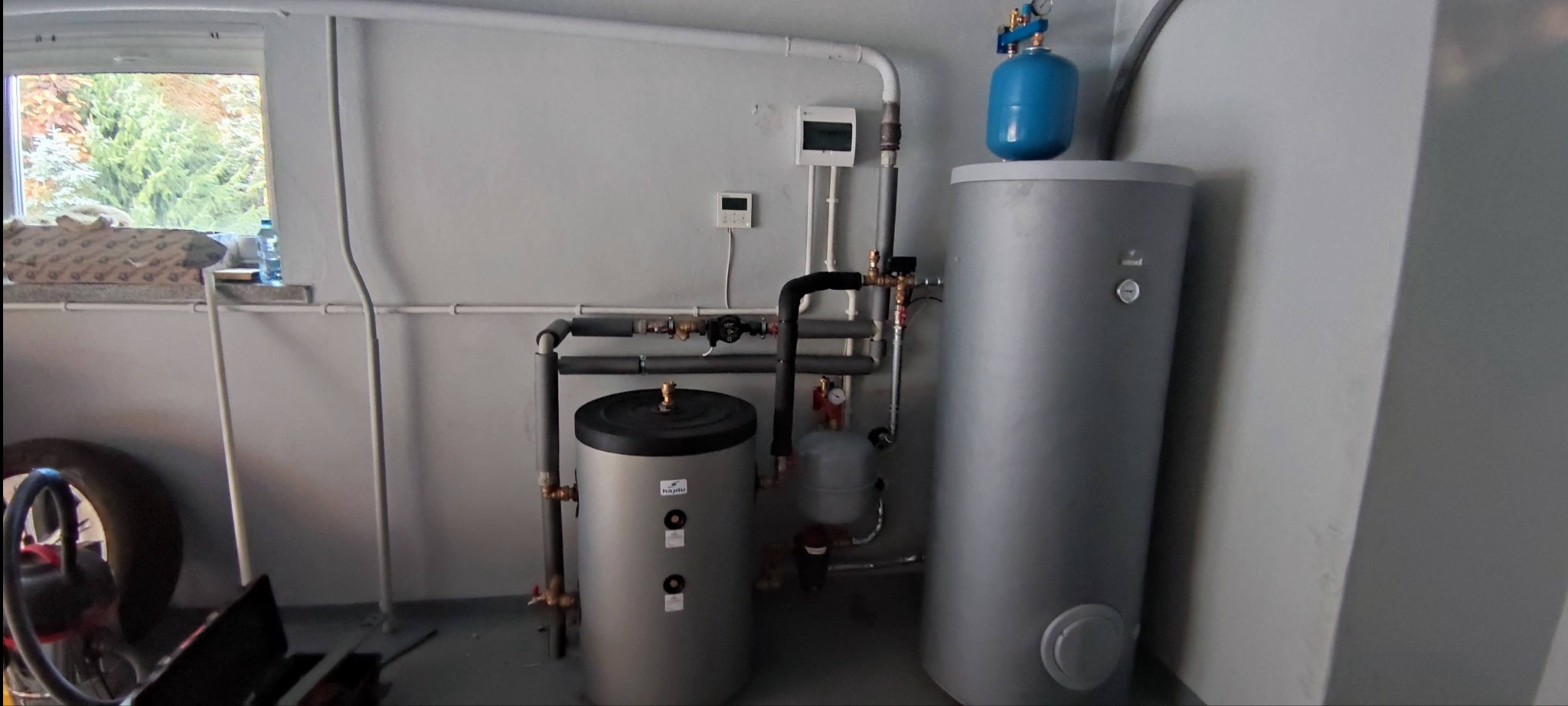 Dwa szare zbiorniki akumulacyjne ciepła w kotłowni, połączone rurami, z niebieskim zbiornikiem wyrównawczym na jednym z nich. Widoczne okno z widokiem na drzewa, regulatory temperatury na ścianie...