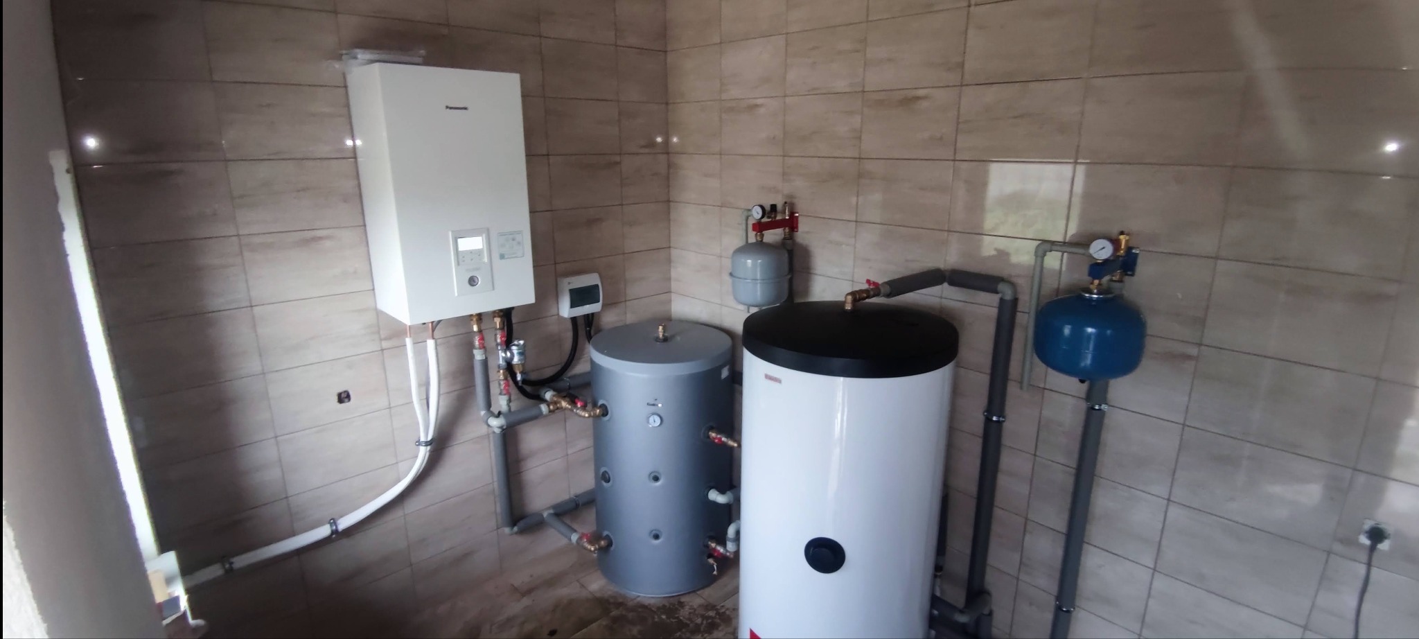 Zainstalowana pompa ciepła powietrze-woda Panasonic z dwoma zbiornikami akumulacyjnymi w pomieszczeniu wyłożonym beżowymi płytkami ceramicznymi.