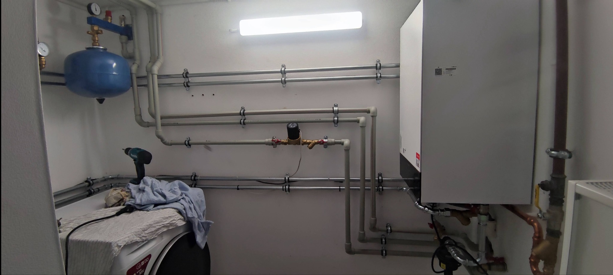 Kompaktowa kotłownia gazowa z białym kotłem wiszącym Viessmann, niebieskim zbiornikiem wyrównawczym i licznymi rurami w małym, oświetlonym pomieszczeniu. Na pralce leżą narzędzia i ubrania.