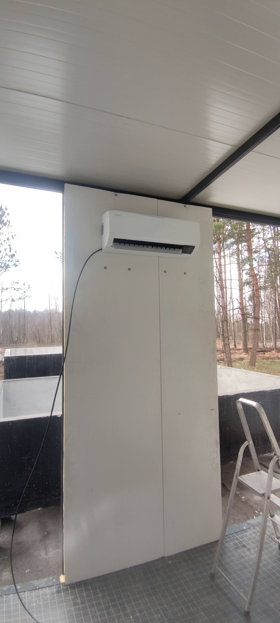 Zamontowany klimatyzator SAMSUNG na białej ścianie w pomieszczeniu z widokiem na drzewa i drabinką.