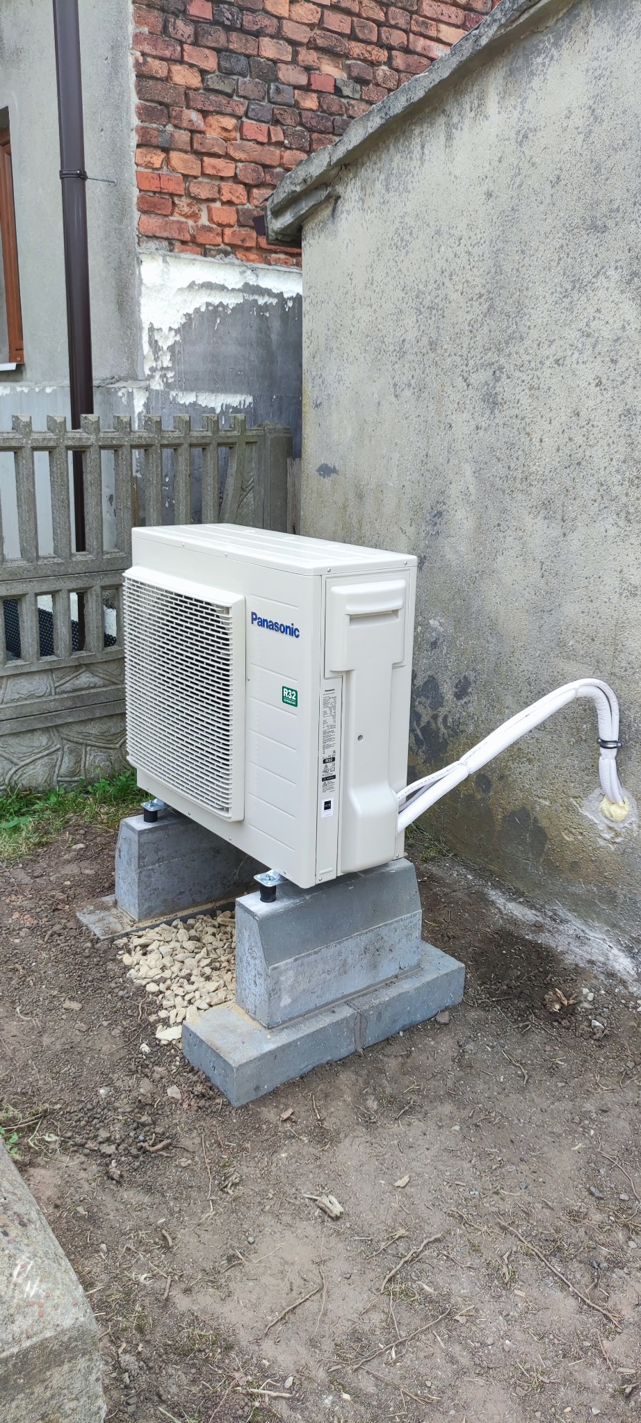 Zewnętrzna jednostka pompy ciepła powietrze-woda Panasonic Aquarea, zamontowana na betonowych blokach fundamentowych, z widocznymi przewodami chłodniczymi w izolacji, na tle ściany budynku z cegły...