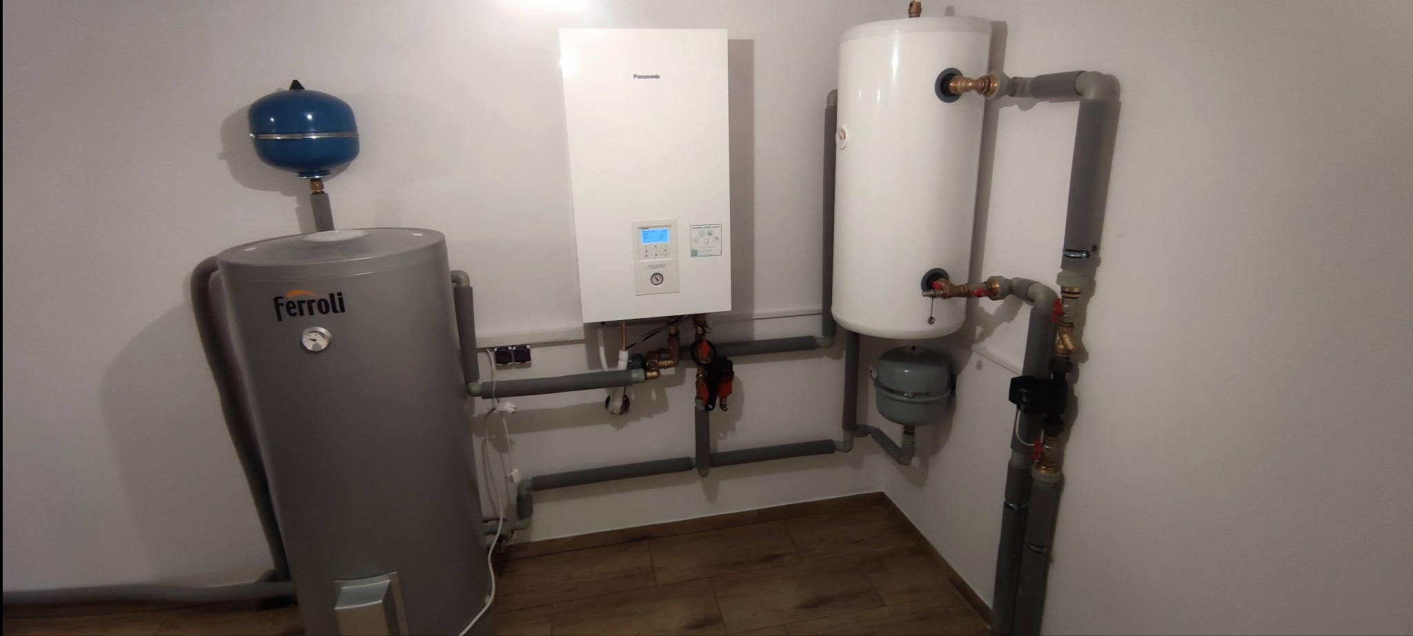 Instalacja grzewcza w kotłowni: pompa ciepła Panasonic, zasobnik Ferolli z niebieskim naczyniem wzbiorczym, biały zbiornik buforowy i szare rury instalacyjne.
