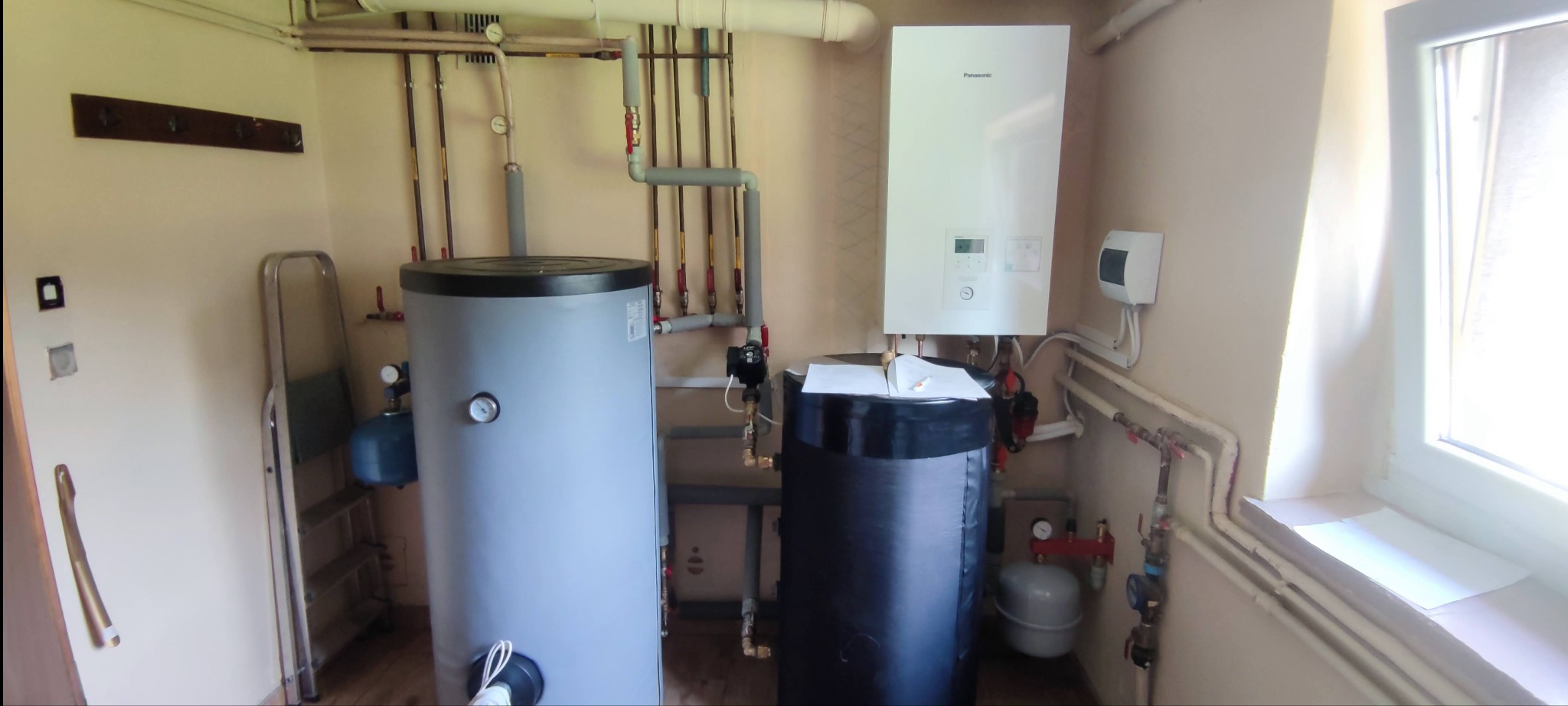 Zainstalowana pompa ciepła powietrze-woda Panasonic w pomieszczeniu technicznym, widoczne dwa zbiorniki akumulacyjne, rury instalacyjne i okno z parapetem.