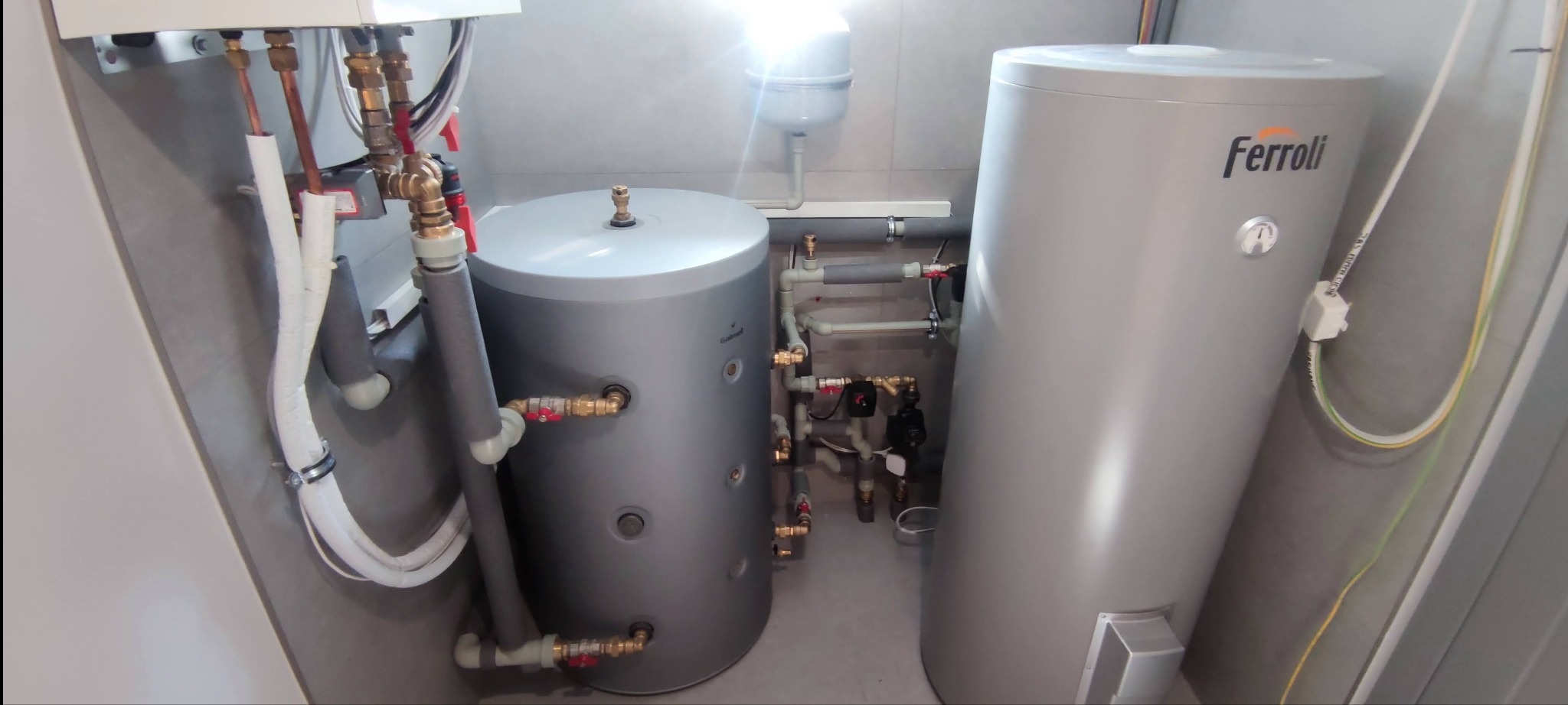 Dwa szare zbiorniki akumulacyjne Ferroli w kotłowni z szarymi płytkami, widoczne rury miedziane, zawory i instalacja hydrauliczna, zasilanie elektryczne poprowadzone w peszlu.