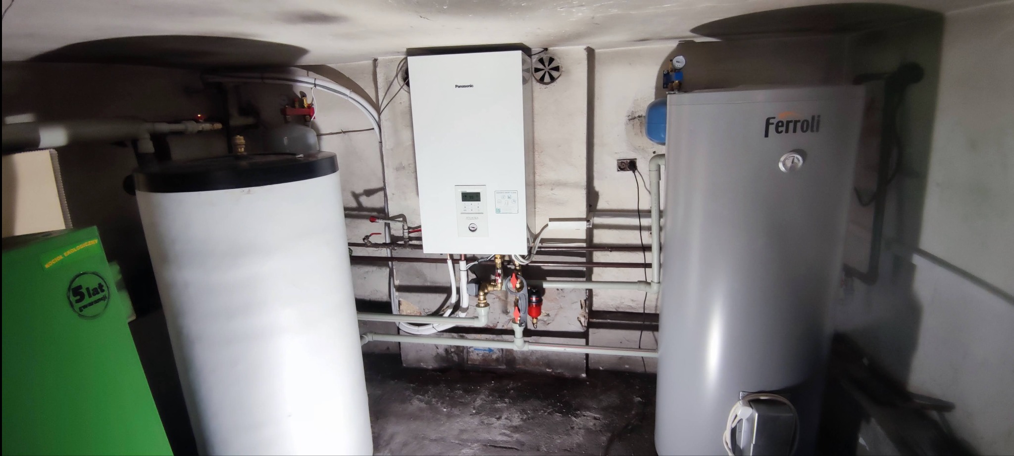 Zainstalowana pompa ciepła Panasonic Aquarea powietrze-woda w piwnicy, obok zbiorników Ferroli, widoczne rury i zawory.