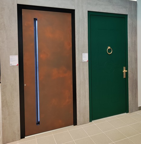 Dwa modele drzwi zewnętrzne: brązowe z podświetlanym pionowym panelem i zielone z kołatką w kształcie pierścienia i złotą klamką, prezentowane w salonie.