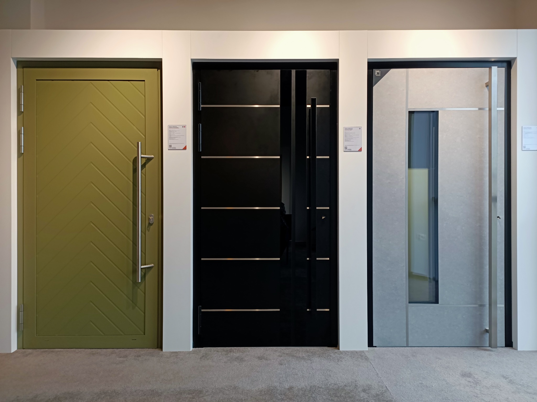 Ekspozycja trzech nowoczesnych drzwi zewnętrznych: oliwkowozielone z ukośnym wzorem, czarne z poziomymi paskami i szare z przeszkleniem, prezentowane na tle białych ścian.