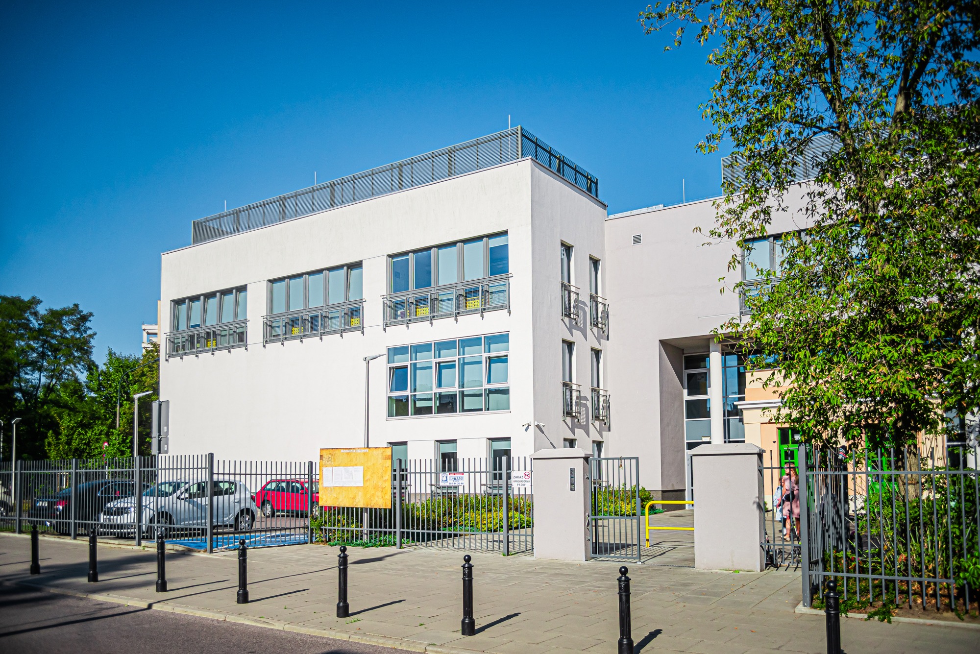 Nowoczesny budynek o białej elewacji z licznymi oknami, widoczny z perspektywy ulicy, ogrodzony metalowym płotem.