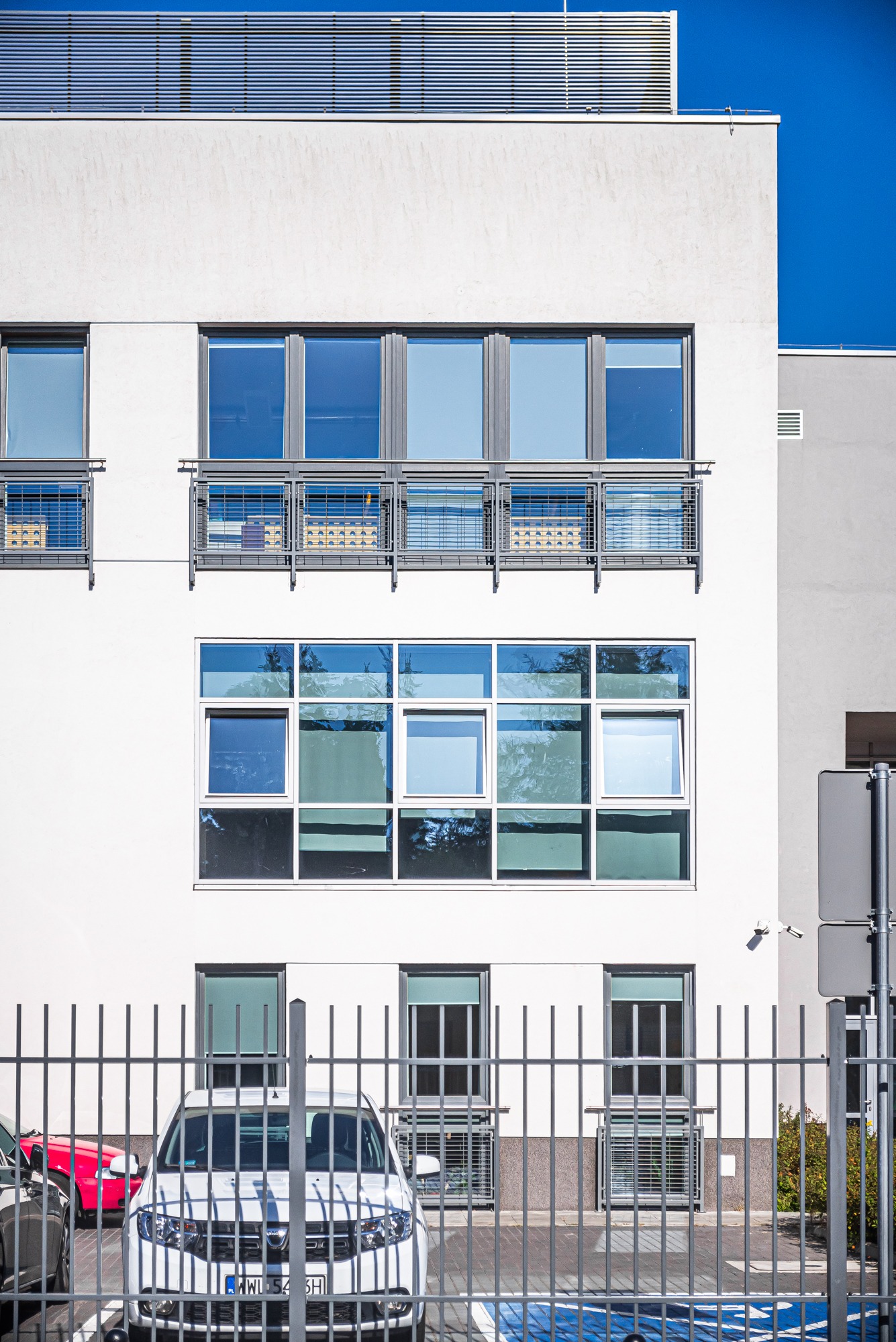 Biały budynek biurowy w Warszawie z nowoczesnymi oknami za metalowym ogrodzeniem, widoczny samochód na parkingu.