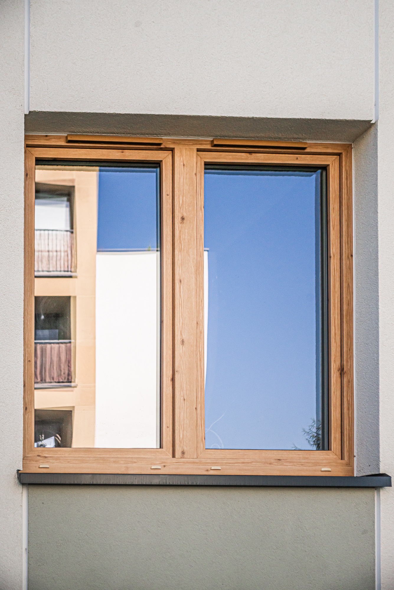 Dwudzielne okno z drewnianą ramą, odbijające błękitne niebo i fragment budynku z balkonami.