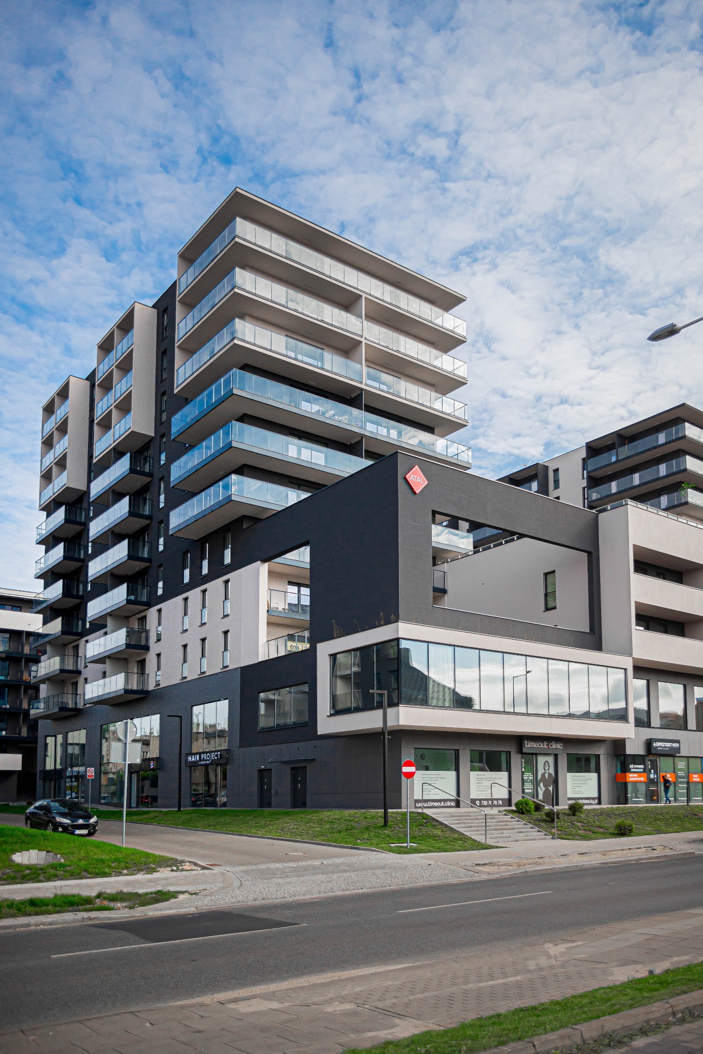 Nowoczesny budynek apartamentowy z balkonami z przeszklonymi balustradami, widoczne duże okna wystawowe na parterze, lokalizacja Łódź-Drewnowska.