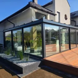 Szklana zabudowa tarasu z czarną ramą, przylegająca do domu z drewnianym tarasem i rabatą z kamykami oraz trawami ozdobnymi. Nowoczesny design.