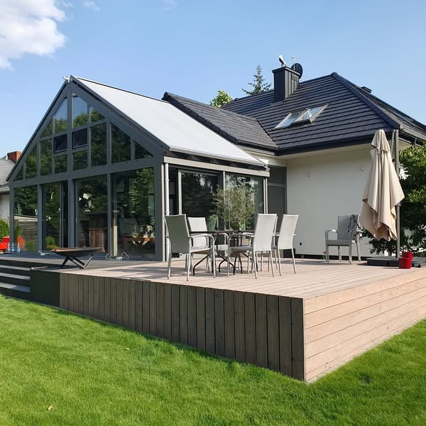 Szklana zabudowa tarasu z metalową ramą, przylegająca do domu. Na tarasie drewniany podest i meble ogrodowe. Widok z zewnątrz, w tle trawnik i fragment domu.