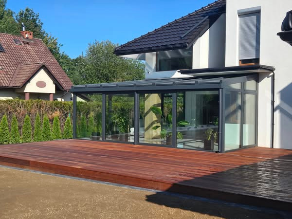 Nowoczesna, przeszklona zabudowa balkonu z metalową ramą i drewnianym tarasem, przylegająca do białego domu z szarą roletą. Widok w słoneczny dzień.