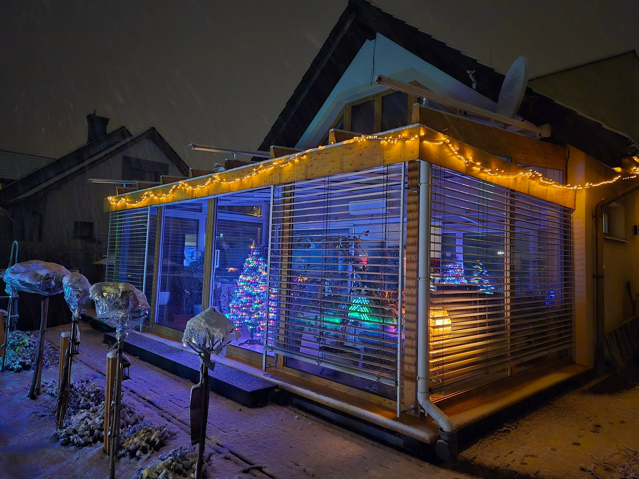 Zimowa zabudowa balkonu z drewnianą konstrukcją, oświetlona girlandą, z widoczną choinką wewnątrz. Śnieg pada, a przed zabudową oszronione rośliny.