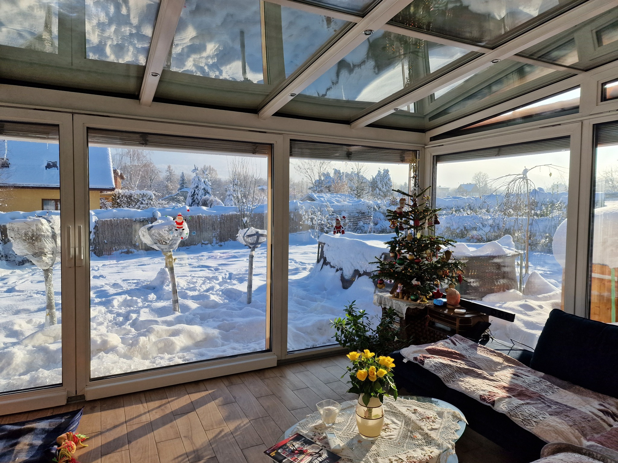 Zimowy ogród zimowy z przeszklonym dachem i dużymi oknami, przez które widać zasypany śniegiem ogród z choinką i dekoracjami świątecznymi, oraz wnętrze z meblami.
