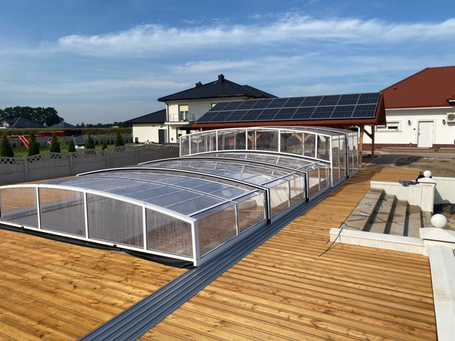 Przesuwna, aluminiowa zabudowa basenu na drewnianym tarasie, z widokiem na domy jednorodzinne i panele słoneczne. Jasne, słoneczne otoczenie.