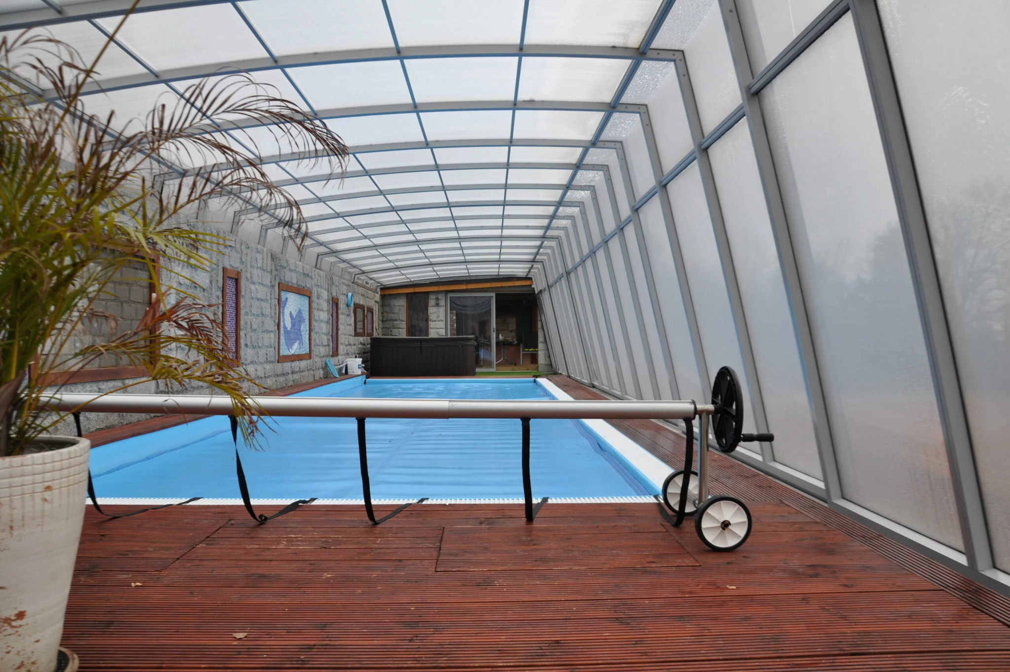 Kryty basen z drewnianym tarasem i rośliną w donicy. Przesuwne zadaszenie basenowe z poliwęglanu. Widoczna część basenu zakryta niebieską plandeką.