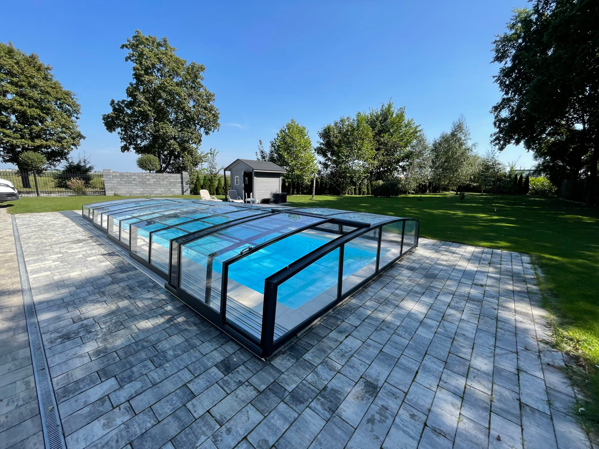Zadaszenie basenu z czarną ramą i przezroczystymi panelami, otoczone szarą kostką brukową na tle zielonego trawnika i drzew w słoneczny dzień.