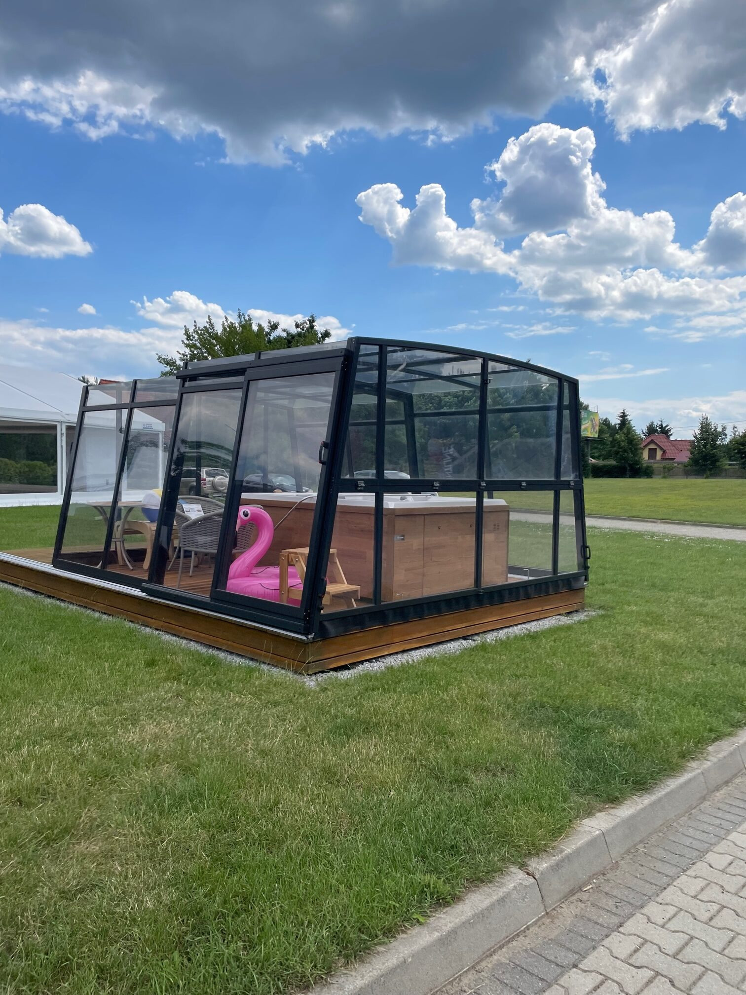 Szklana zabudowa tarasu z czarną ramą, na drewnianym podeście, z jacuzzi i dmuchanym flamingiem w środku, na tle zielonego trawnika i błękitnego nieba.