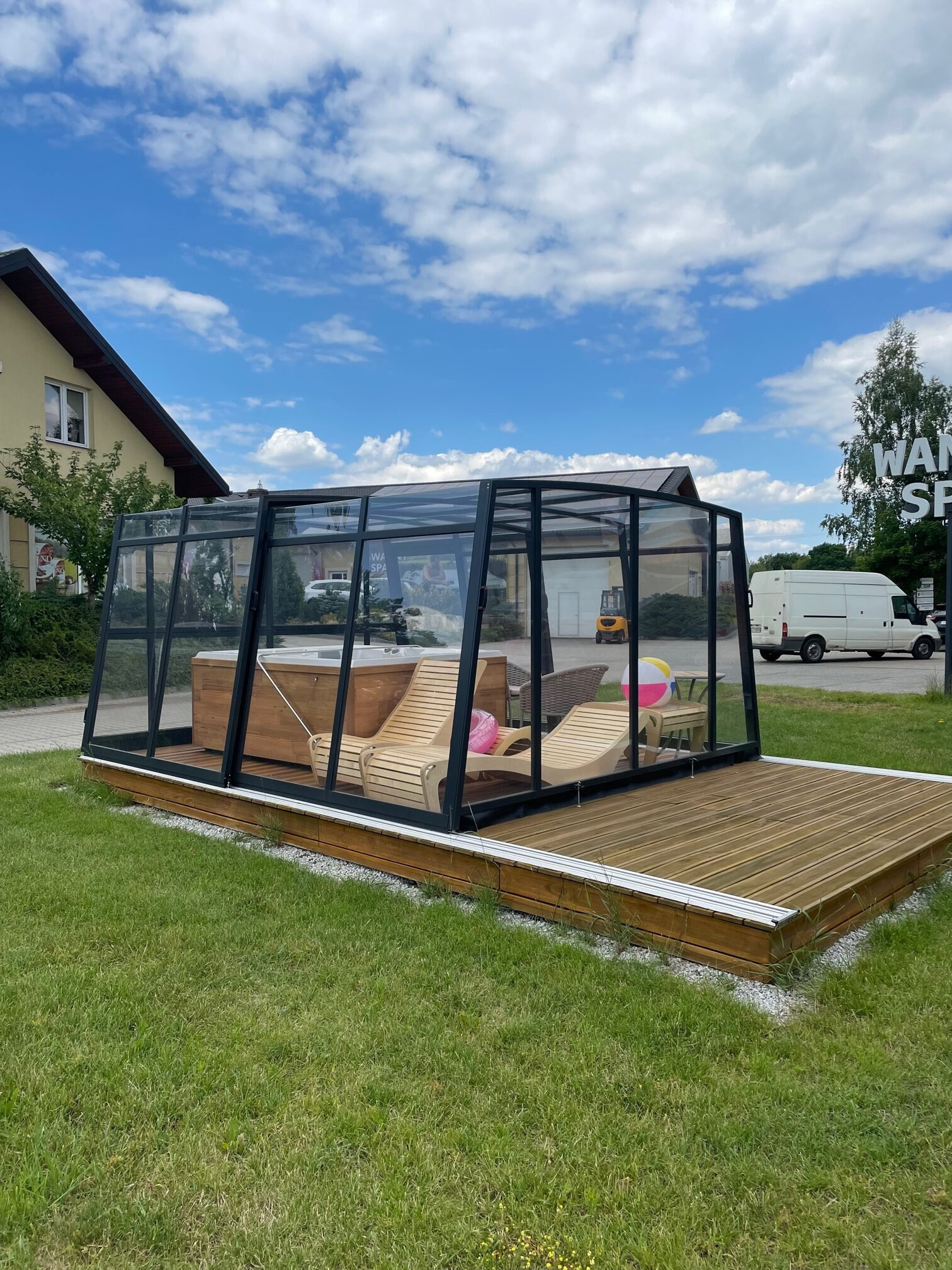 Szklana konstrukcja z jacuzzi i leżakami na drewnianym podeście, otoczona zielenią w Bujakowie. Nowoczesna zabudowa z widocznymi detalami wykończenia.