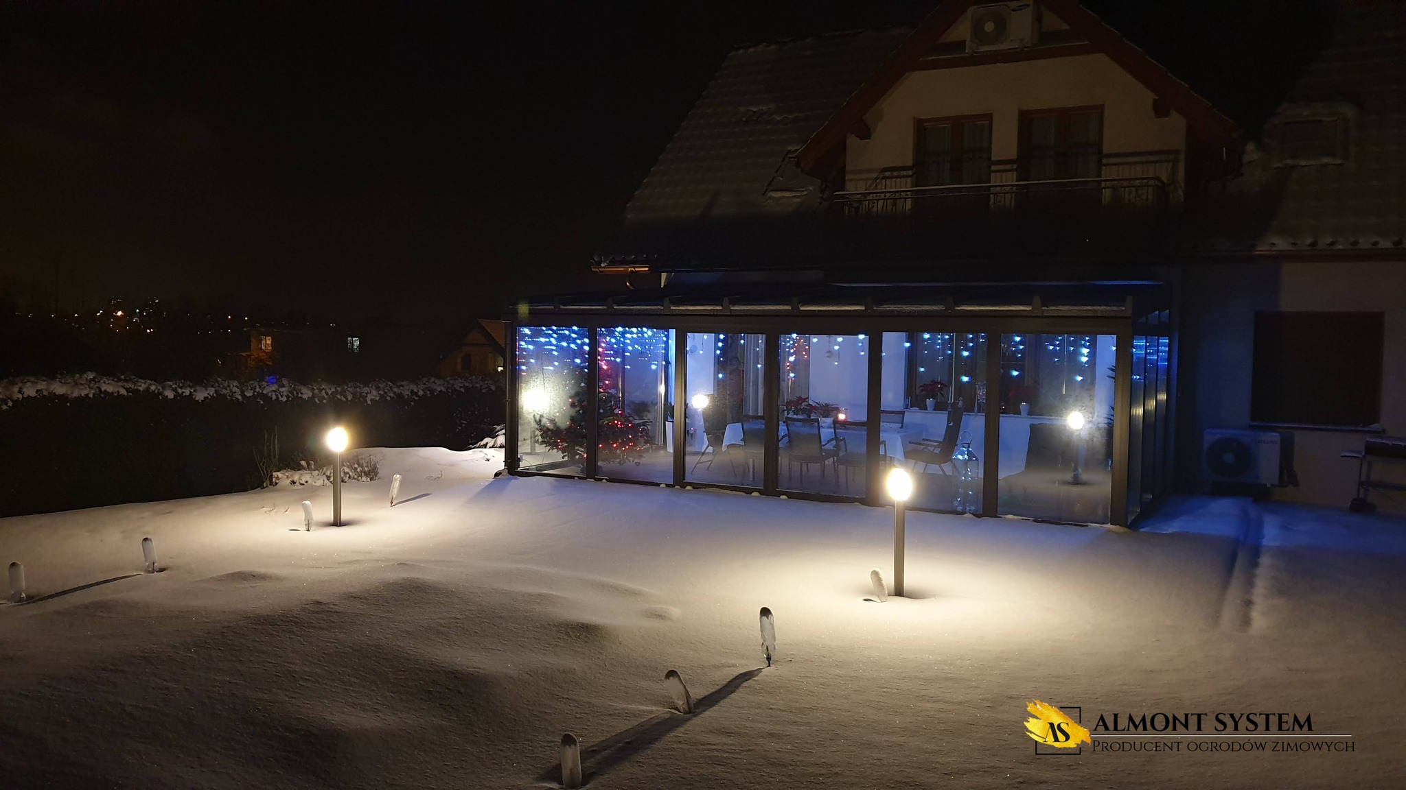 Zimowa zabudowa balkonu z metalową konstrukcją i oświetleniem LED. Wnętrze ozdobione świątecznie, widoczne z zewnątrz przez szklane ściany. Śnieżny ogród oświetlony lampami.