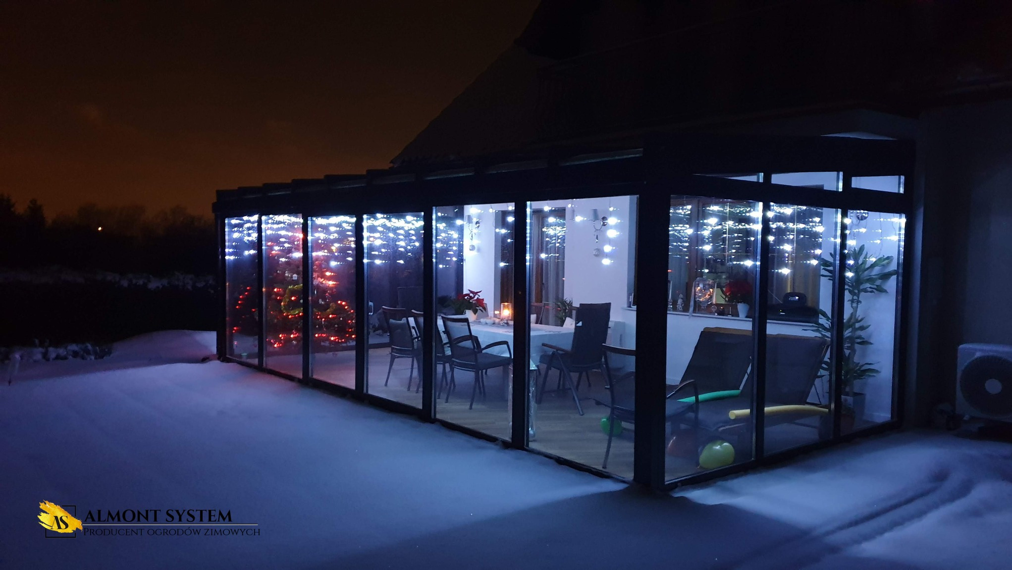 Zimowy ogród z metalową konstrukcją i szklanymi ścianami, oświetlony świątecznymi lampkami. Wewnątrz stół z krzesłami i choinka. Na zewnątrz śnieg.
